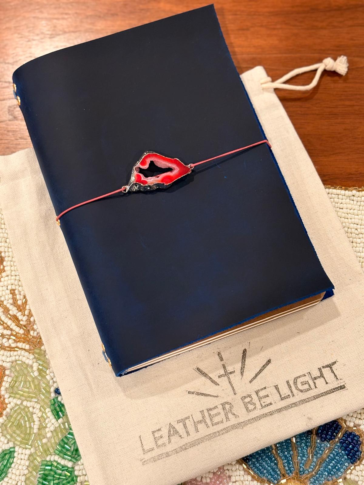 Blue leather-bound notebook with a red geode charm on a white sack with 'Leather Be Light' branding.