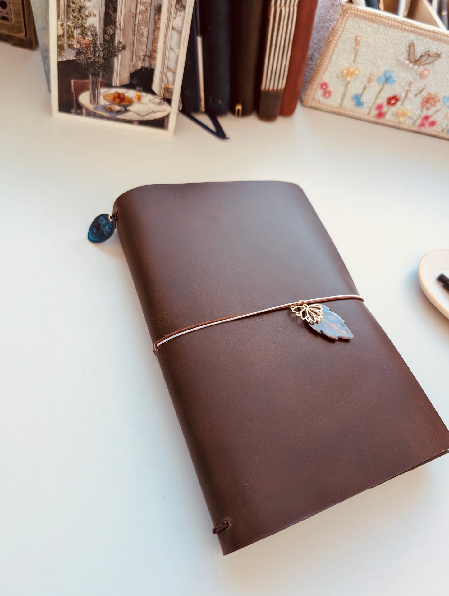 Brown leather notebook with elastic closure and decorative charms on a white surface